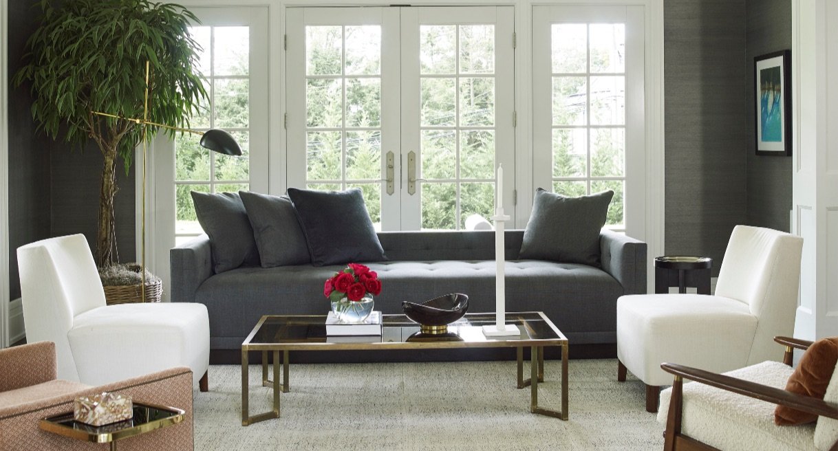 Living room with charcoal grasscloth walls, a dark tufted sofa, white slipper chairs and a brass coffee table