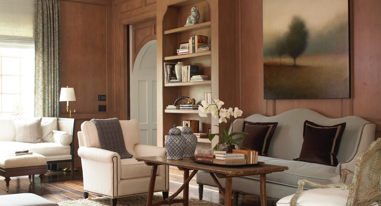 Warm wood paneled library with a camelback sofa, nailhead club chairs, built-in bookshelves and a landscape painting